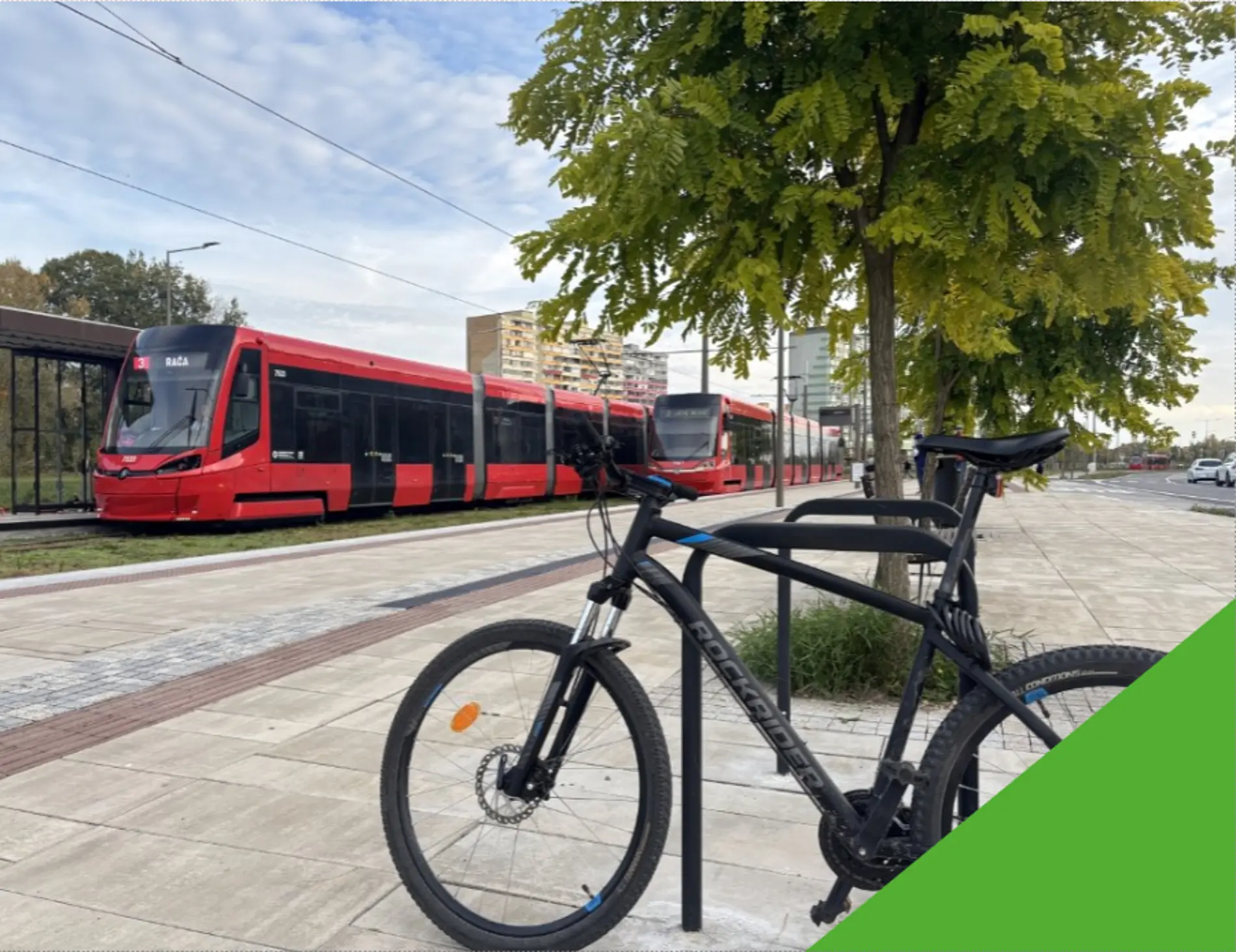 A black bicycle is locked to a metal stand near a tree on a paved sidewalk. In the background, a long red tram is stopped on tracks, with apartment buildings and greenery visible under a partly cloudy sky.