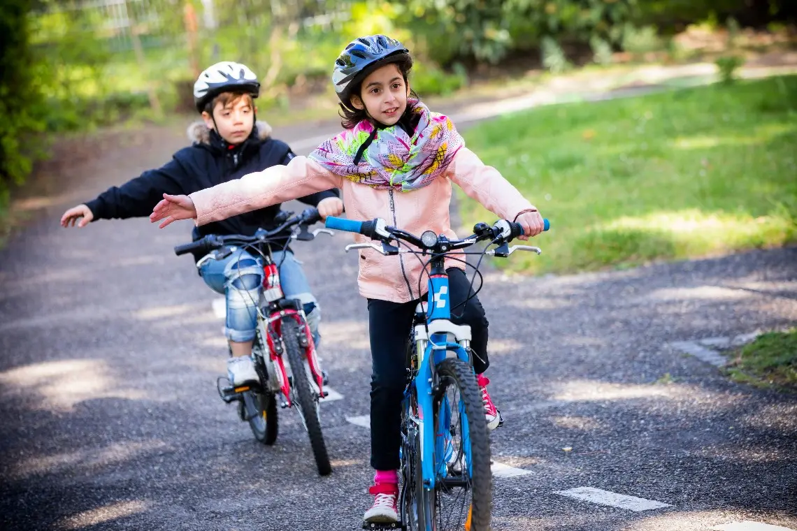 Zwei Kinder fahren Fahrrad und üben das Rechtsabbiegen. Sie halten die rechte Hand seitlich ausgestreckt während sie mit dem Fahrrad fahren. Beide tragen Helm.
