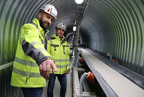 Zwei Mitarbeitende des Bad Ischler Trockenmörtelwerks stehen in gelben Warnwesten neben der neu errichtetetn Förderbandanlage.