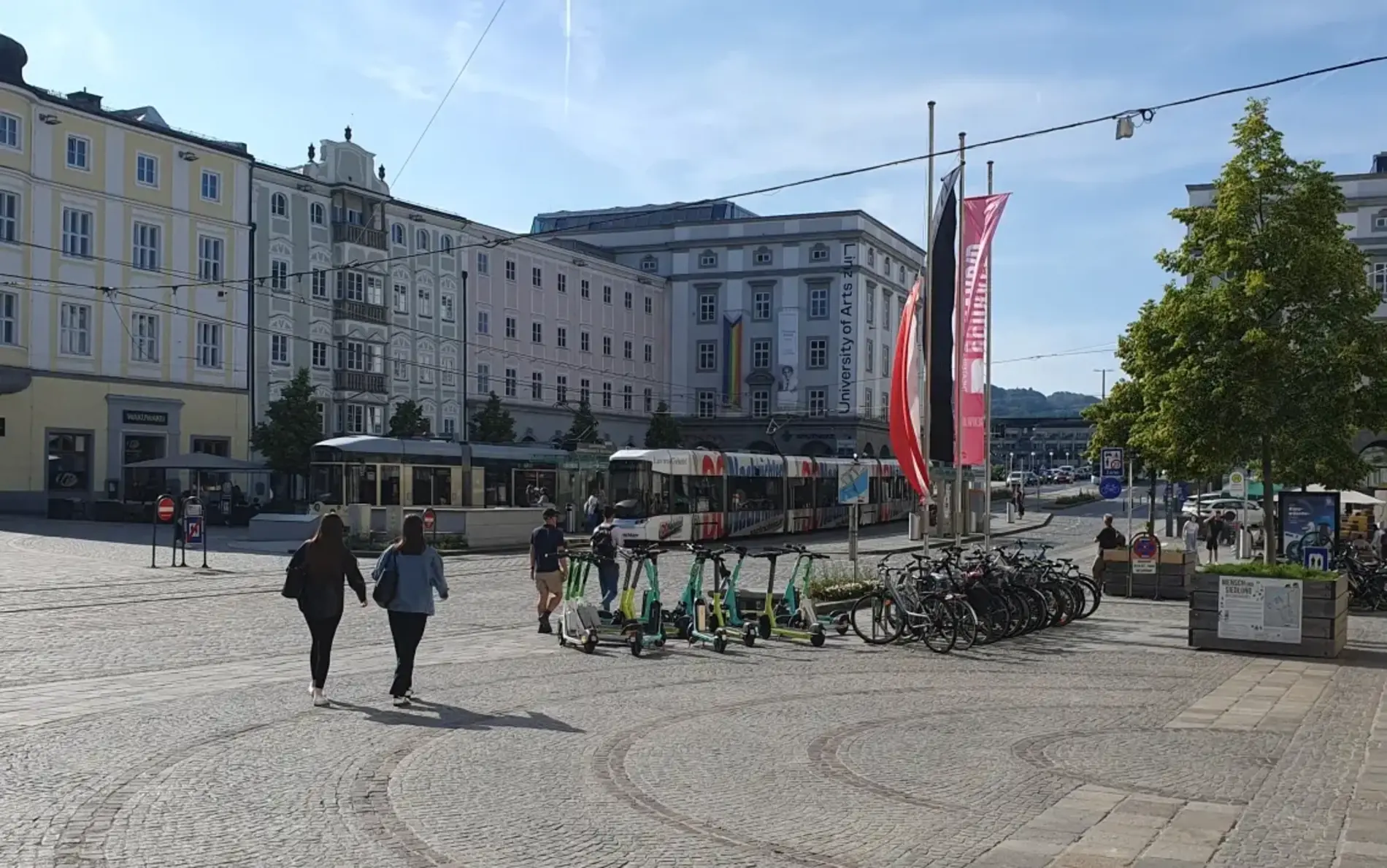 A cobblestone square in a historic city center with several people walking and a row of parked bicycles and e-scooters in the foreground. A tram is visible in the background near pastel-colored buildings, and two tall red flags stand beside a tree under a clear blue sky.