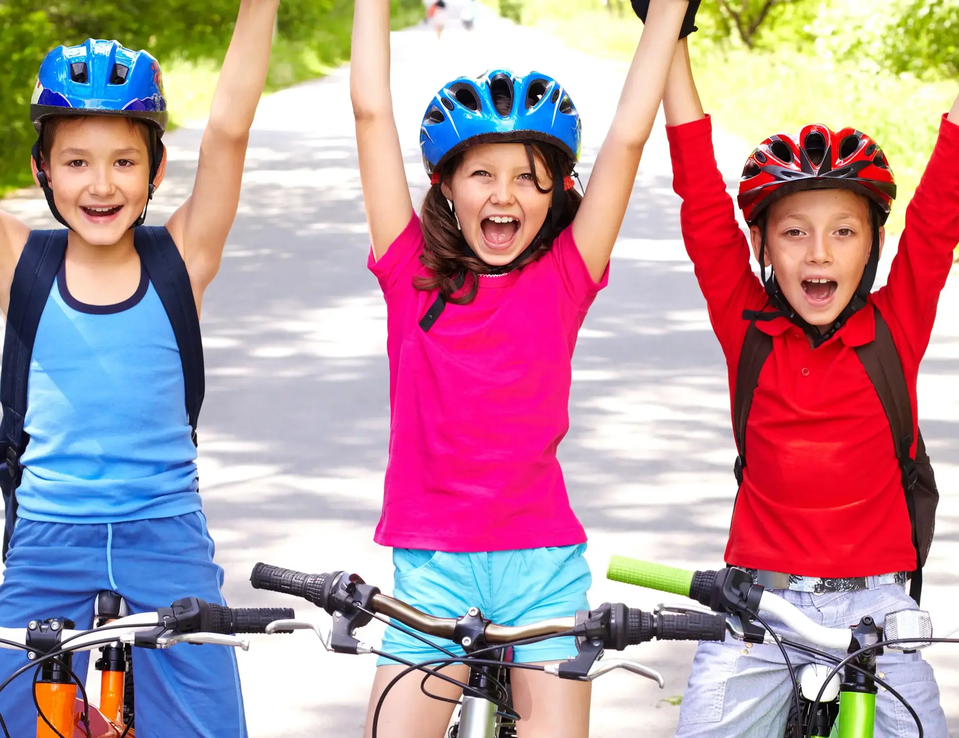 Drei Kinder sitzen am Fahrrad, die Füße abgestellt. Sie halten die Hände in die Höhe und rufen freudig. Alle drei tragen Helm