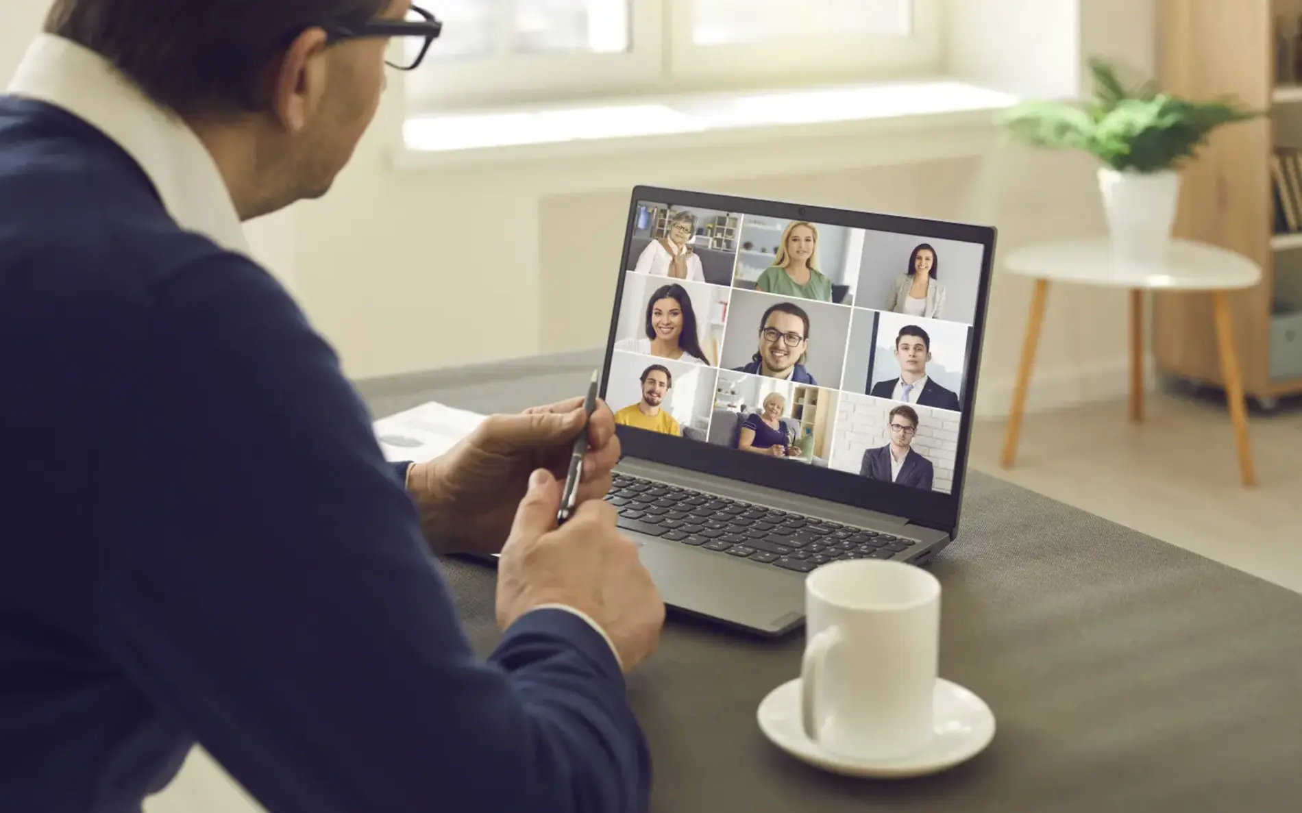 Mann mit Brille sitzt vor Laptop und hält Videokonferenz. Am Bildschirm verschiedene Gesichter