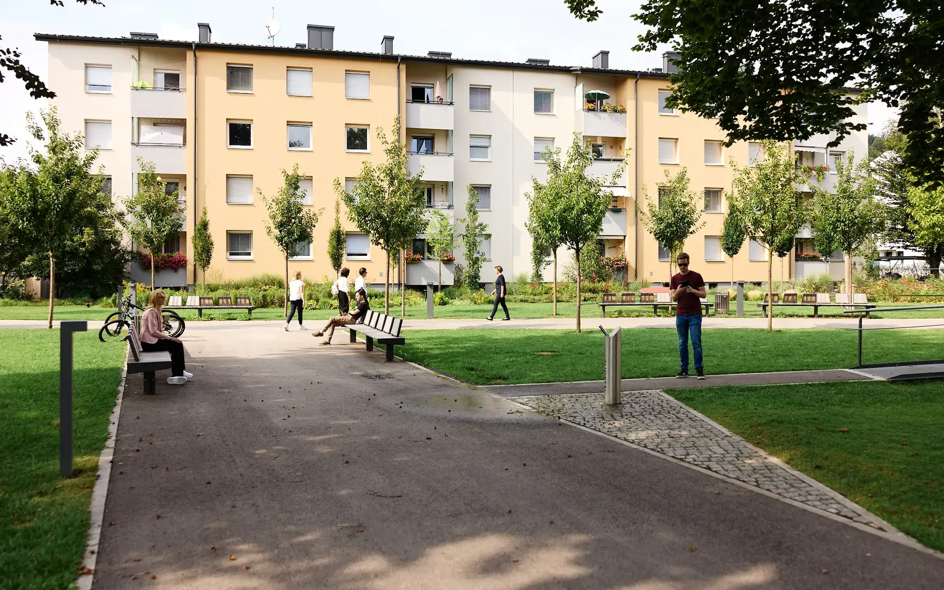 Ein breiter Gehweg durch eine Siedlung. Links und rechts ist der Weg begrünt - es scheint ein Innenhof zu sein.