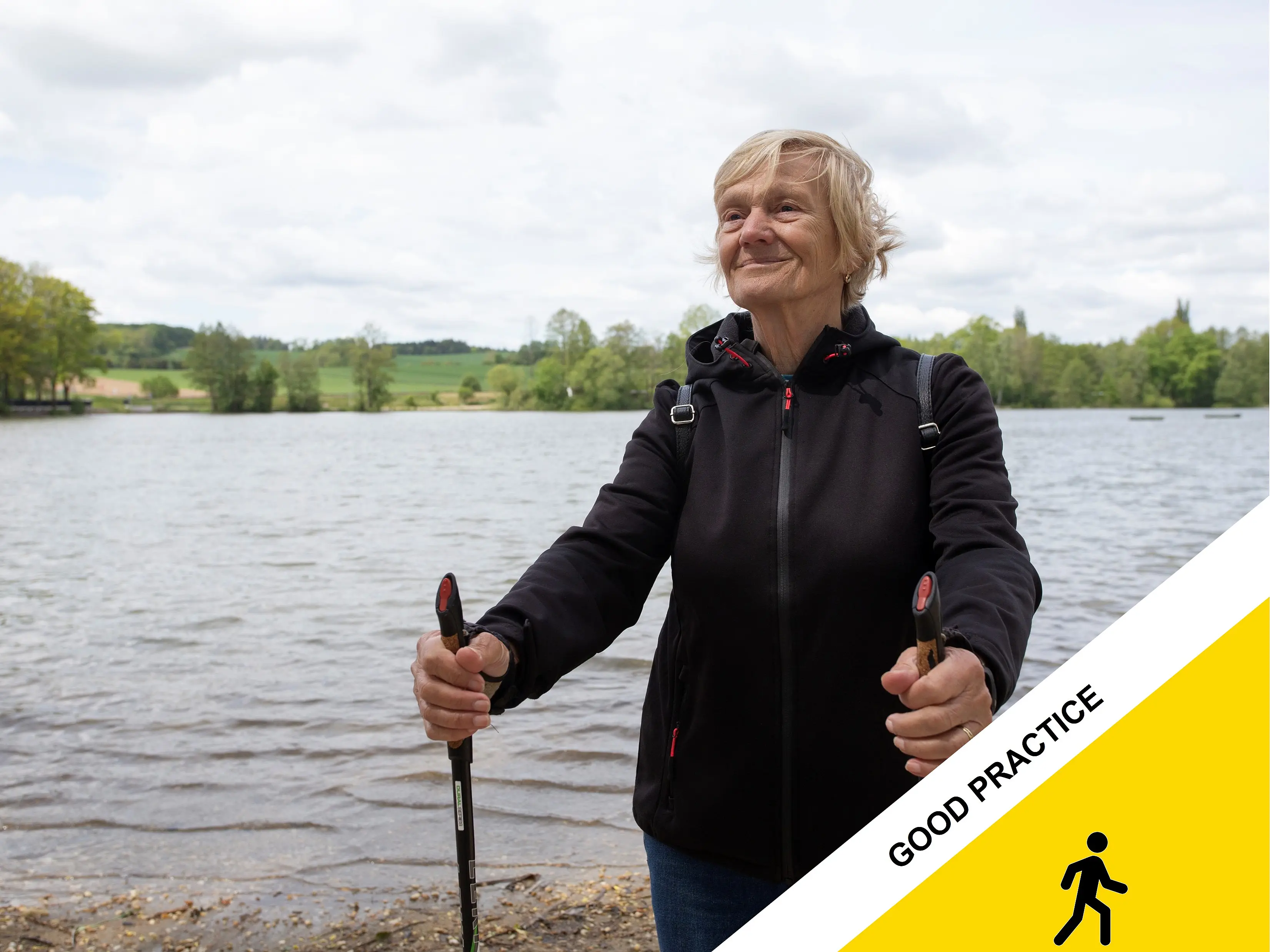 An elderly woman walking by the lake with Nordic walking poles.  