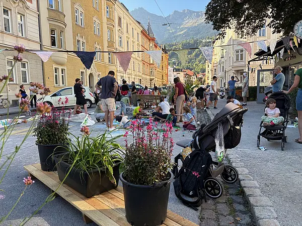 Auf einer gesperrten Straße stehen Bänke, Blumenkübel, viele Menschen spielen, essen und trinken. Weit im Hintergrund ein Bergpanorama. links und rechts Stadthäuser in gelb