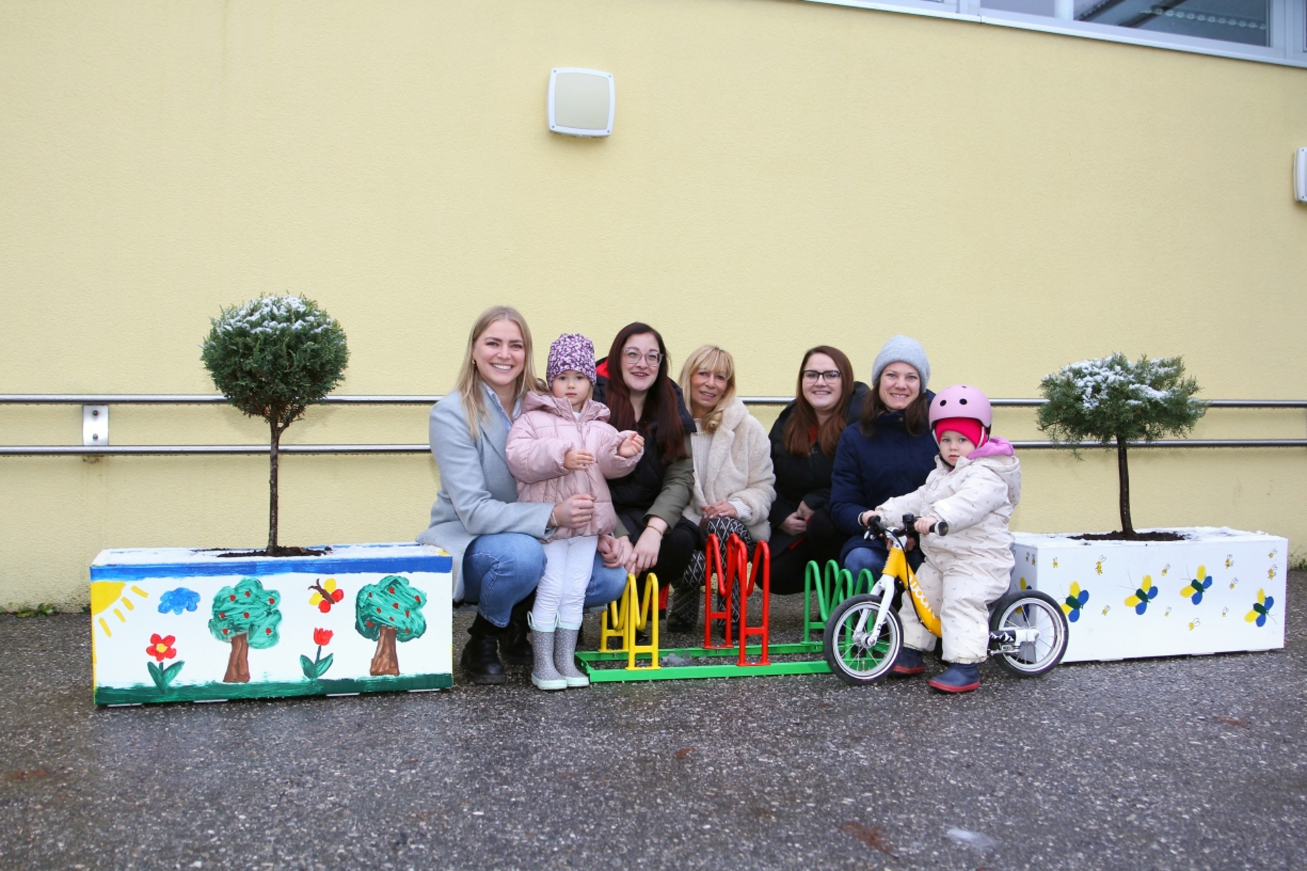 Kinder und Kindergartenpädagoginnen posieren vor breiten Blumentrögen, die bemalt sind. Sie stehen nahe des Eingangs des Kindergartens.