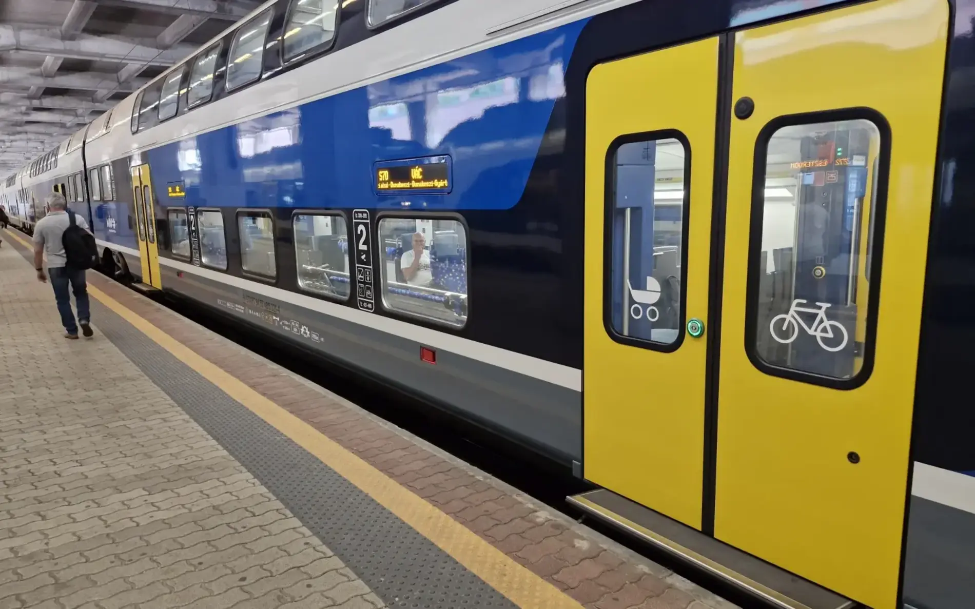 Ein blau-gelber Doppelstockzug steht am Bahnsteig in einem überdachten Bahnhof. Die geöffnete Tür zeigt Symbole für Kinderwagen und Fahrräder, und ein Mensch geht am Zug entlang.