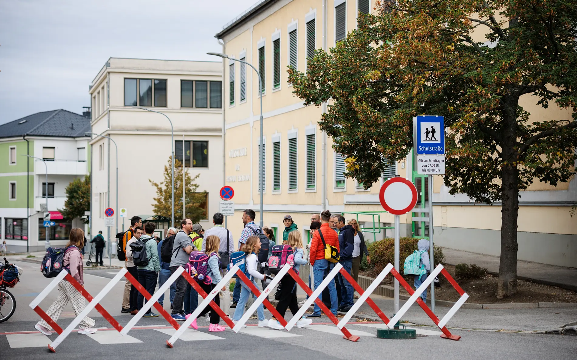 Eine Straße gesperrt mit Scherengitter, dahinter gehen Kinder frei auf der Straße. Am Rand ist das Schulstraßenschild zu sehen. Im Hintegrund die Schule