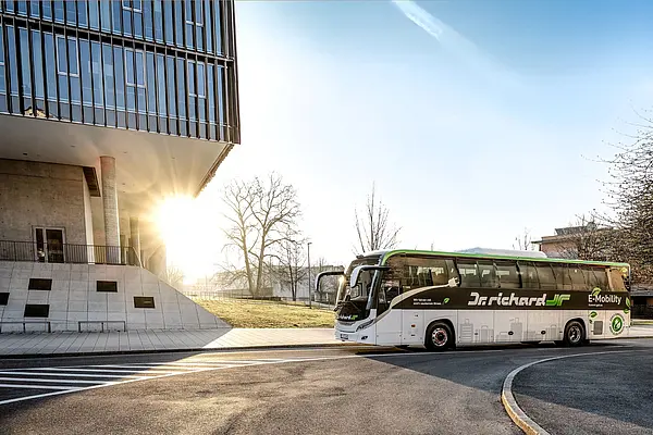 rechts: elektrischer Reisebus vor blauem Himmel, links: hohes Gebäude mit Glasfront auf Betonstütze