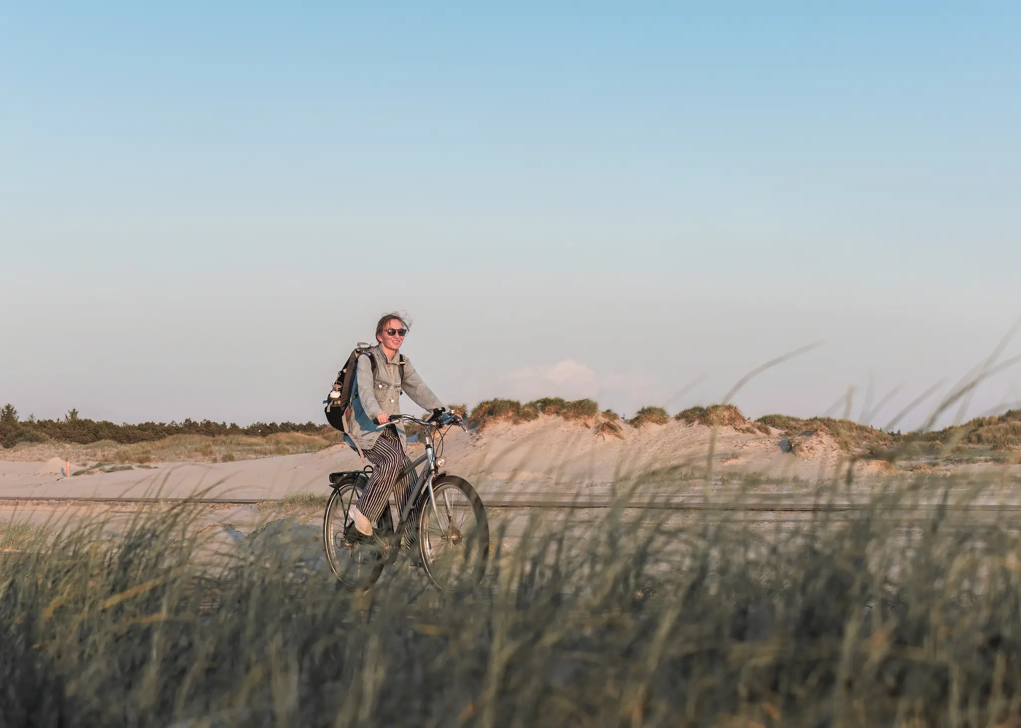 junge Frau auf dem Fahrrad, im Vordergrund hohe Gräser, im Hintergrund Sanddünen. 