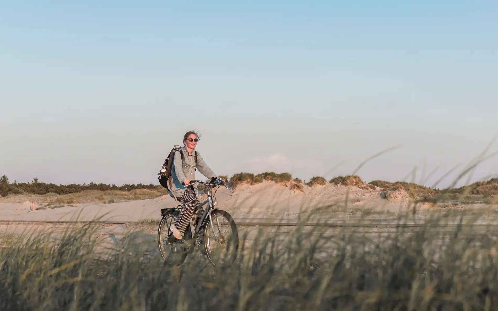 junge Frau auf dem Fahrrad, im Vordergrund hohe Gräser, im Hintergrund Sanddünen. 