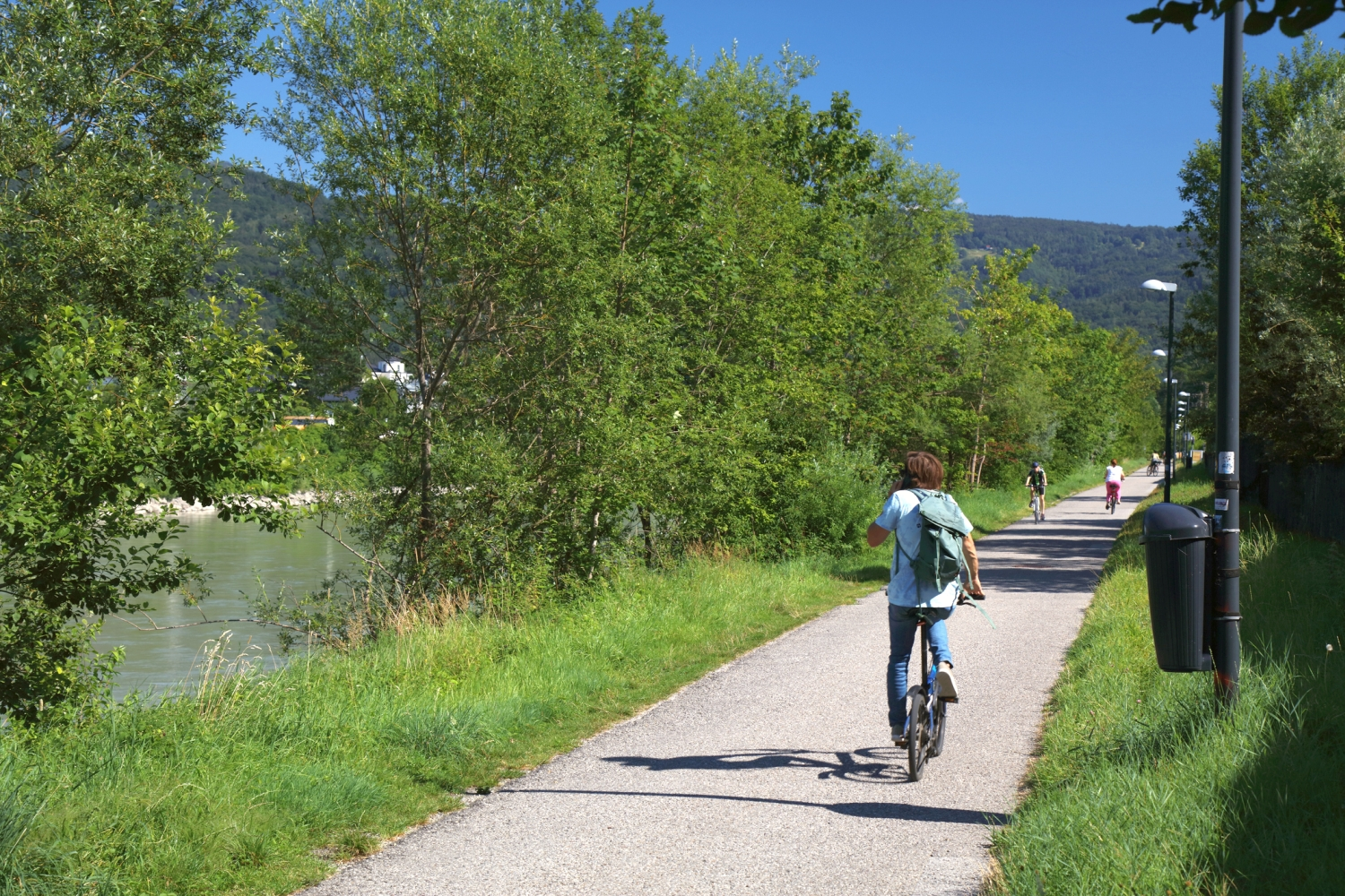 Eine Radfahrerin auf einem Faltrad am Tauernradweg von hinten. Links fließt ein Fluss, direkt neben dem Weg ist alles begrünt und mit Laternen ausgestattet.
