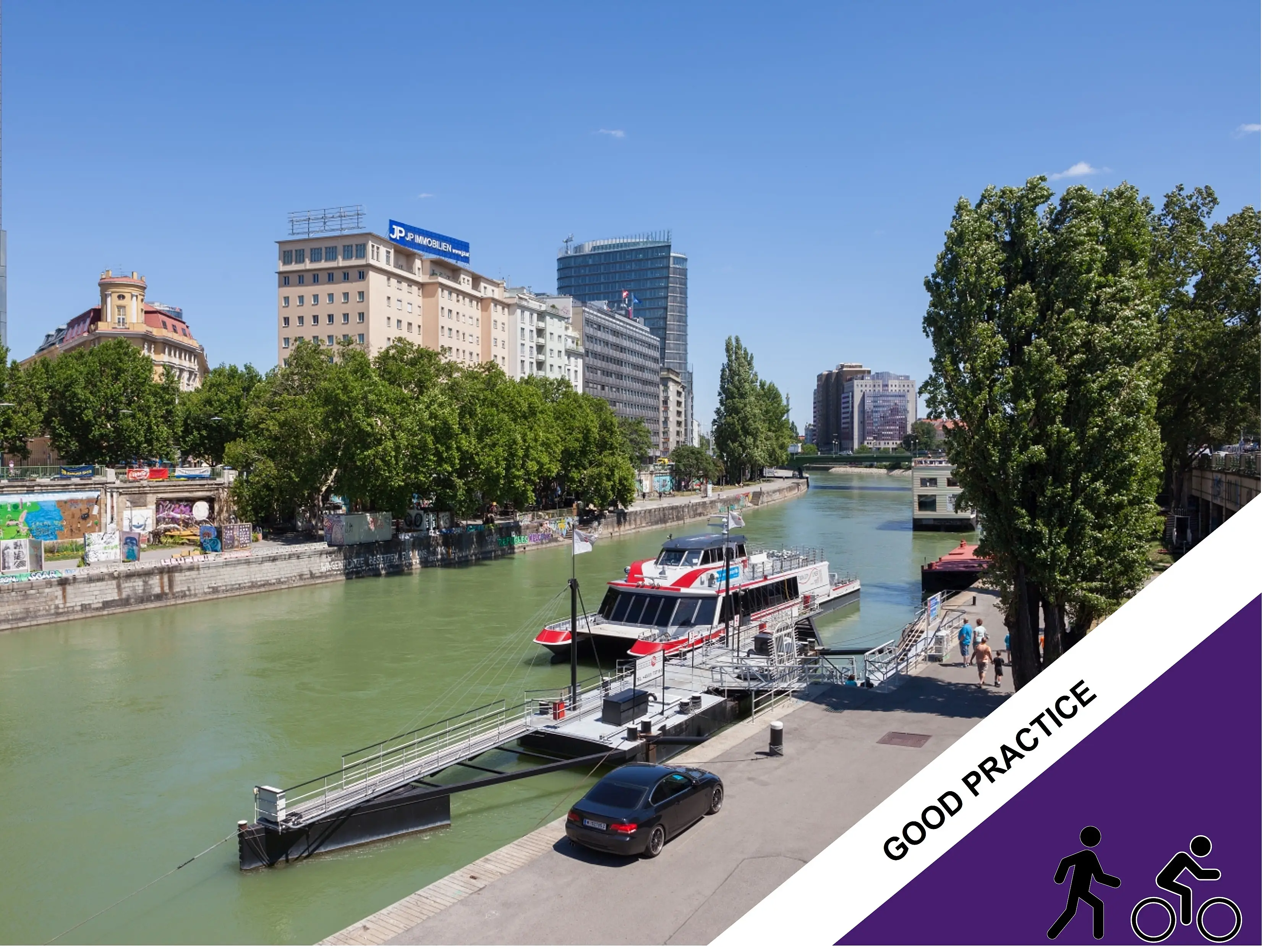 Twin City Liner passenger vessel moored at a dock on the Danube Canal in Vienna, with trees and city buildings lining the waterfront under a clear blue sky.