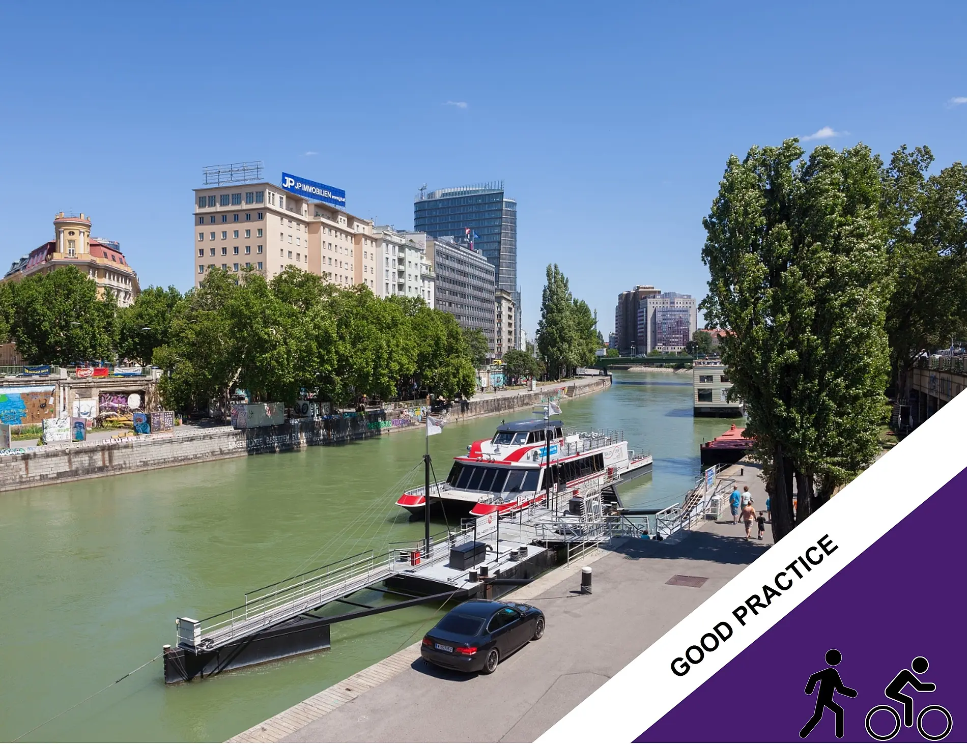 Twin City Liner passenger vessel moored at a dock on the Danube Canal in Vienna, with trees and city buildings lining the waterfront under a clear blue sky.