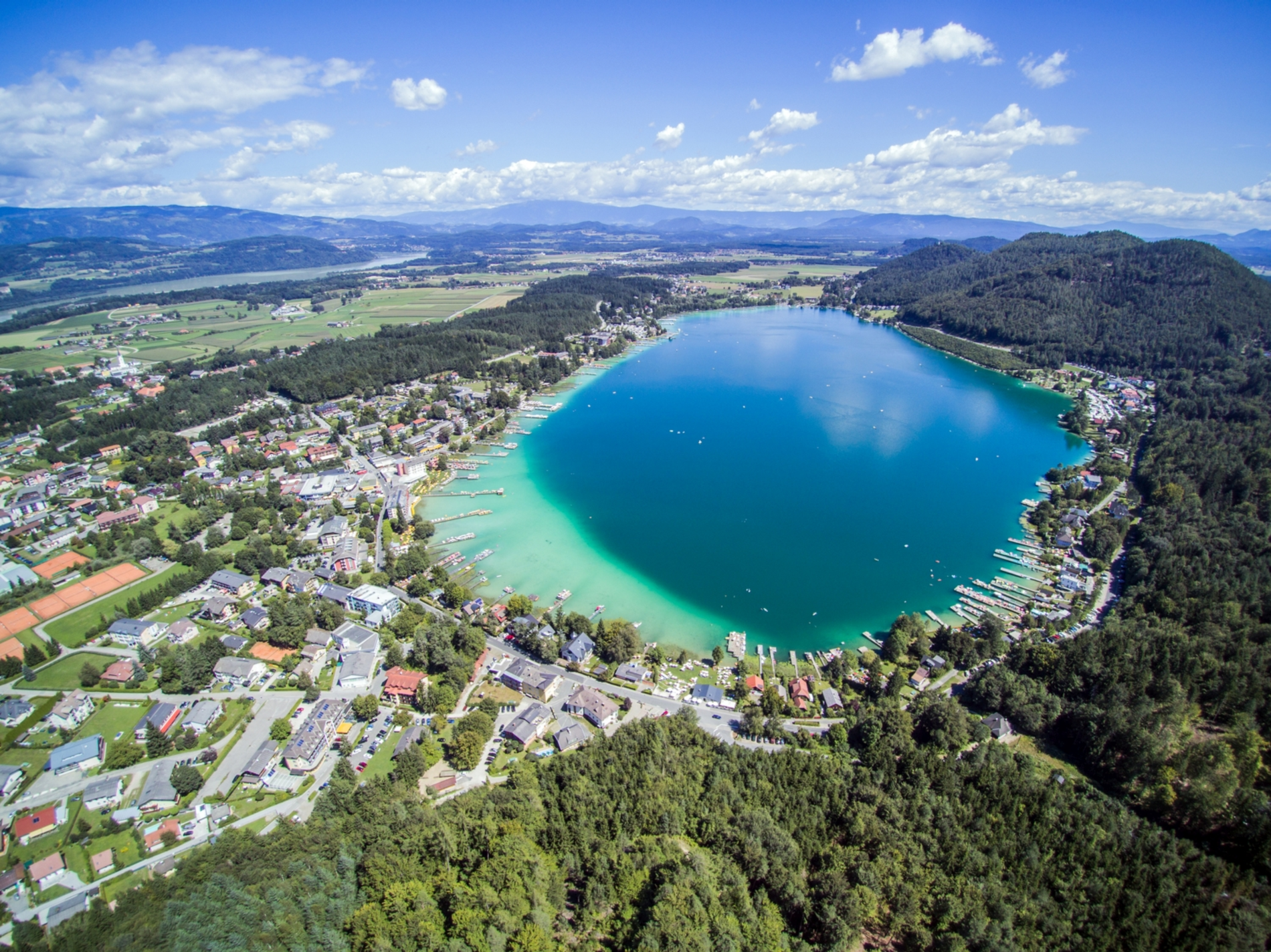 Sicht von oben auf den Klopeiner See. Einige Stege ragen in den See hinein. Eine Uferseite ist voll mit Häusern, die andere Seite vor allem von Wald umgeben.