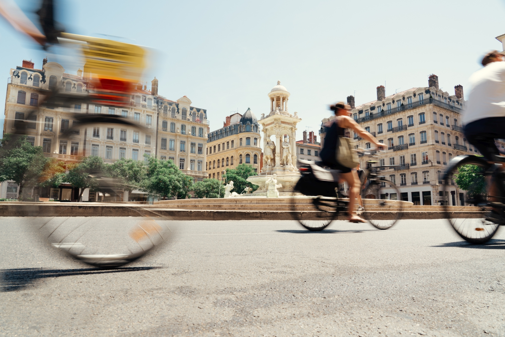 Drei Radfahrende in Bewegung fahren an einem runden Platz in Lyon vorbei. Es scheint die Sonne und die Radfahrenden tragen Sommerkleidung.