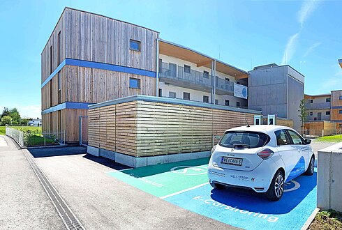 Ein Wohngebäude aus Holz, davor zwei Parkplätze für E-Autos. Ein Auto parkt darauf mit der Aufschrift "E-Carsharing"