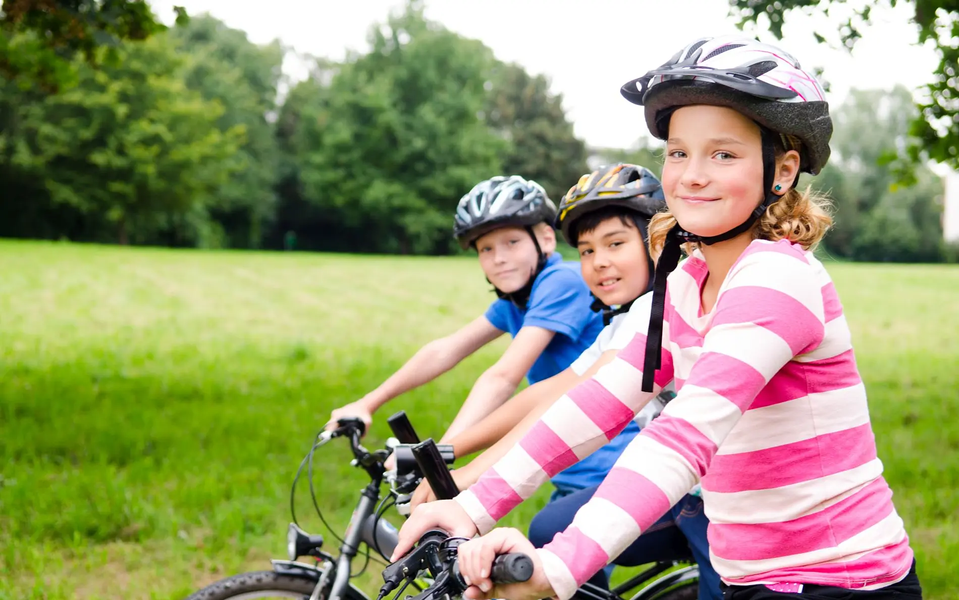 3 Kinder schräg hintereinander auf dem Fahrrad und mit Helm. Lächeln in die Kamera