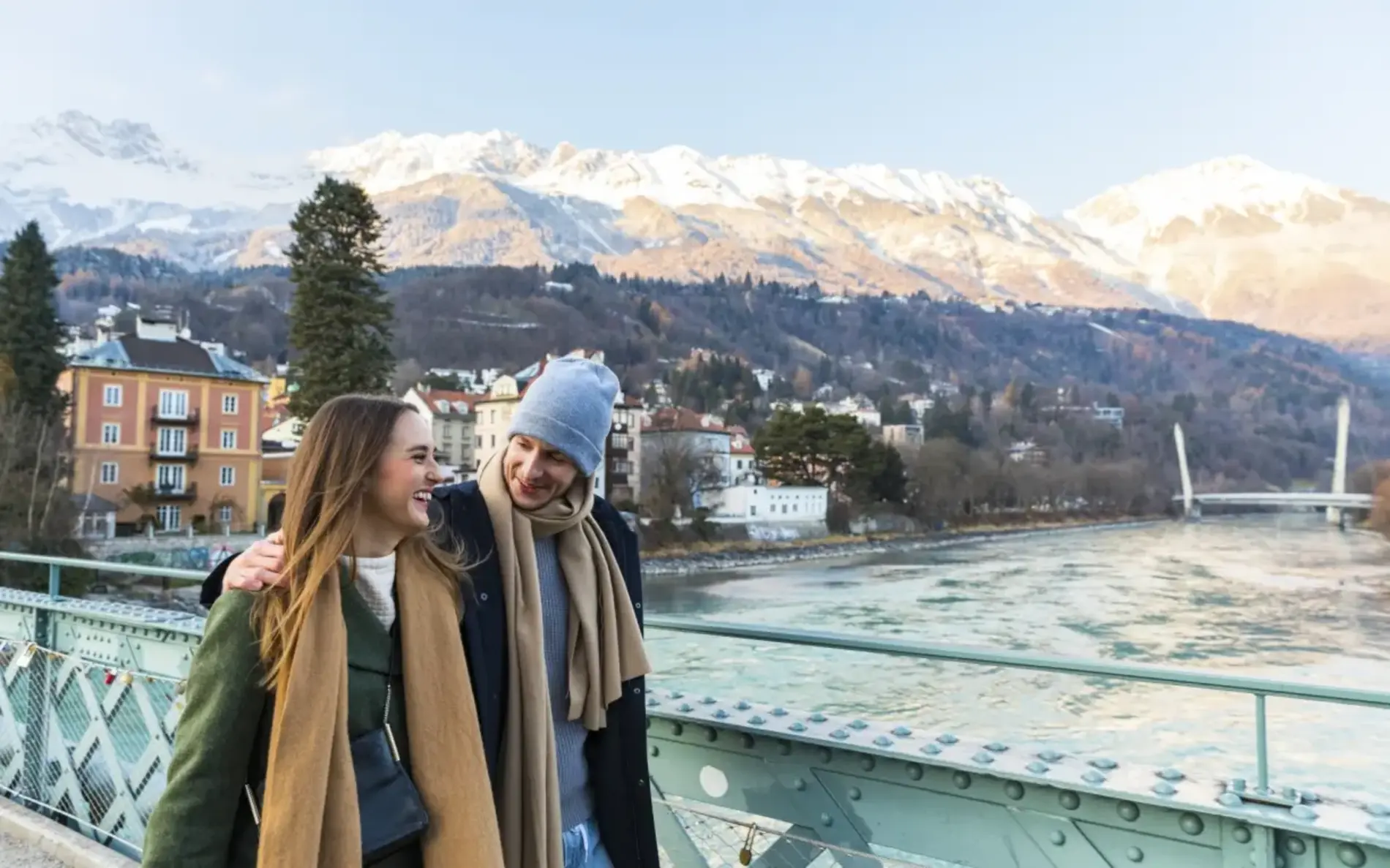 Junges Paar mit Haube und Schal geht über eine Brücke, die über den Inn führt. Im Hintergrund Berge mit Schnee und Sonne