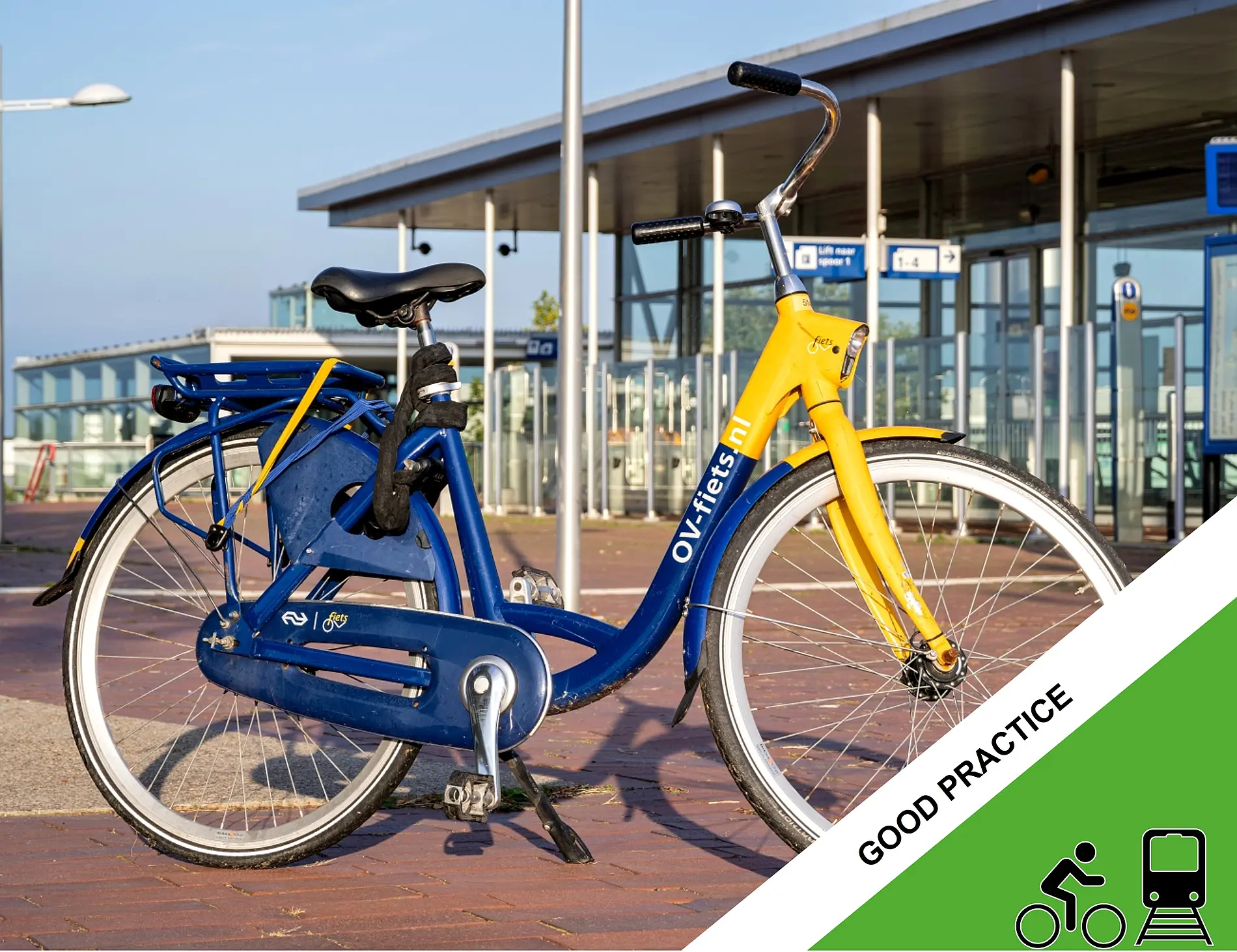 A blue and yellow OV-fiets shared bicycle with the text “OV-fiets.nl” on the frame is parked outside a modern public transport station with glass walls and platform signage in the background.
