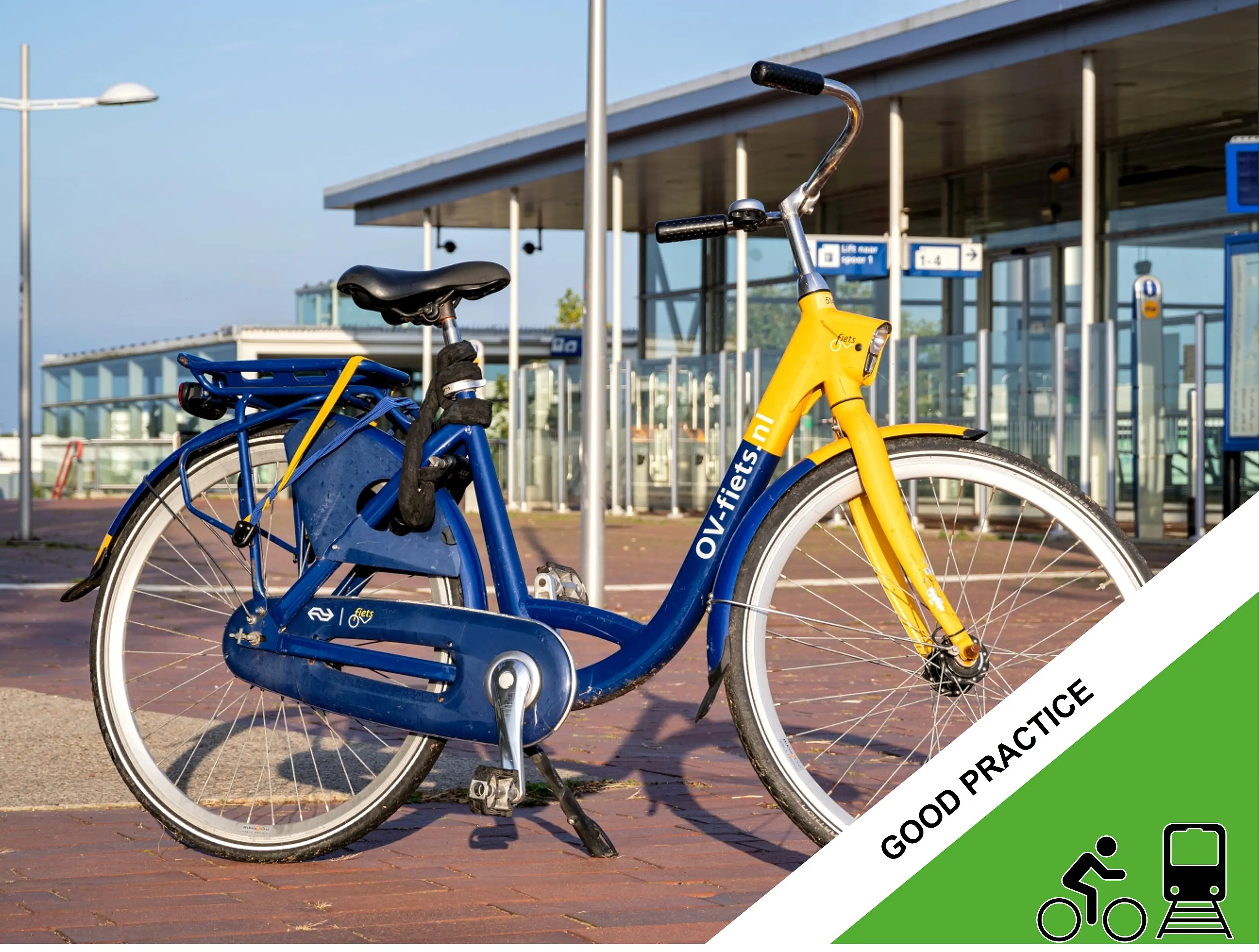 A blue and yellow OV-fiets shared bicycle with the text “OV-fiets.nl” on the frame is parked outside a modern public transport station with glass walls and platform signage in the background.