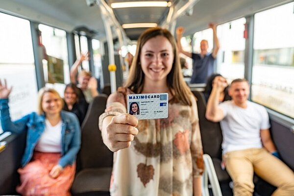 Mehrere Personen sitzen im Bus, in der Mitte steht eine Frau und hält grinsend ein Öffi-Ticket in die Kamera