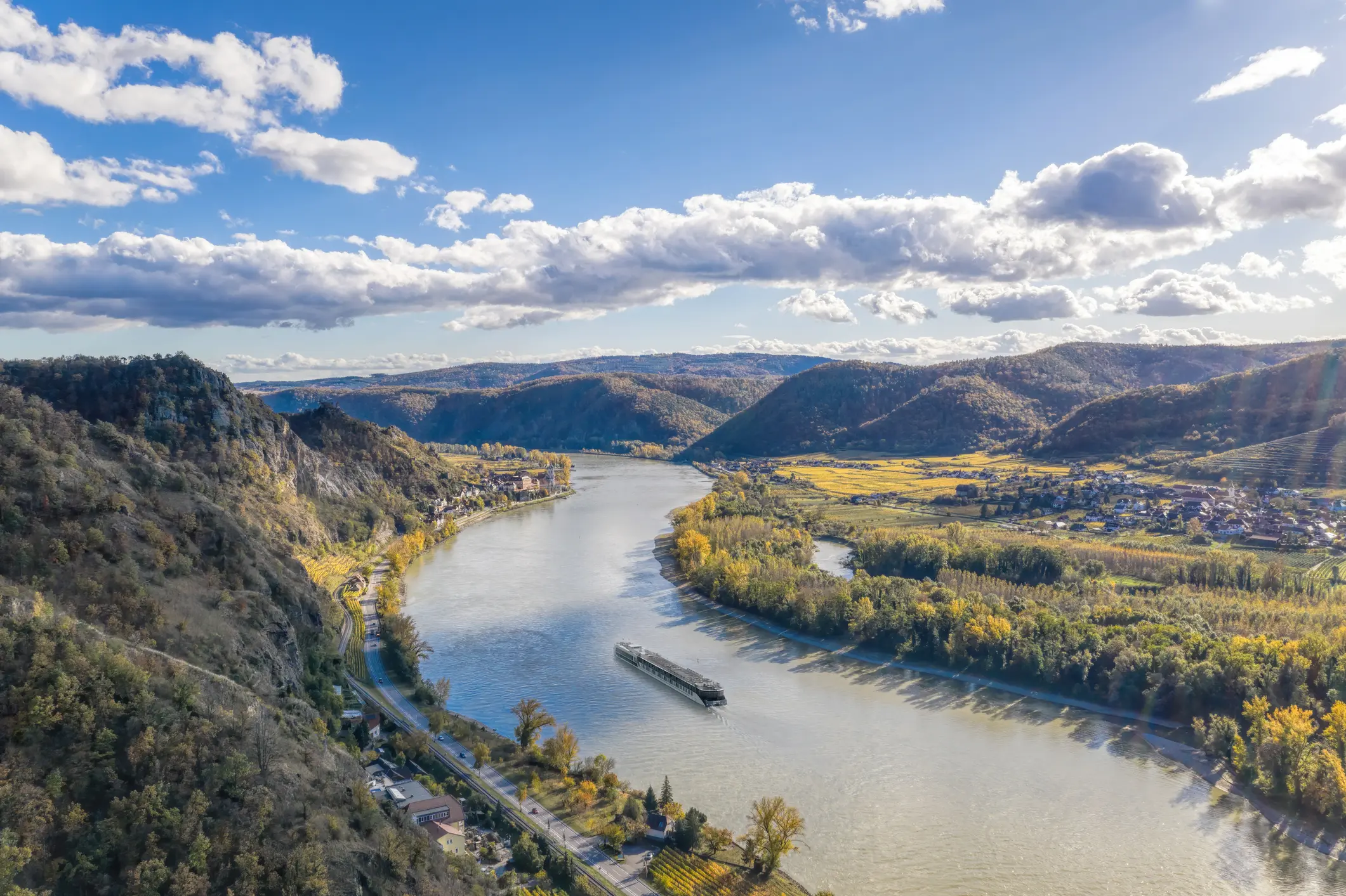 Die Donau von oben, links Gebirge, rechts eine kleine Gemeinde und viel Grün. Am Fluss fährt eine Fähre
