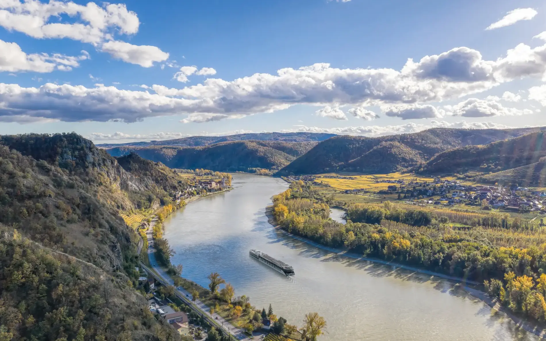 Die Donau von oben, links Gebirge, rechts eine kleine Gemeinde und viel Grün. Am Fluss fährt eine Fähre
